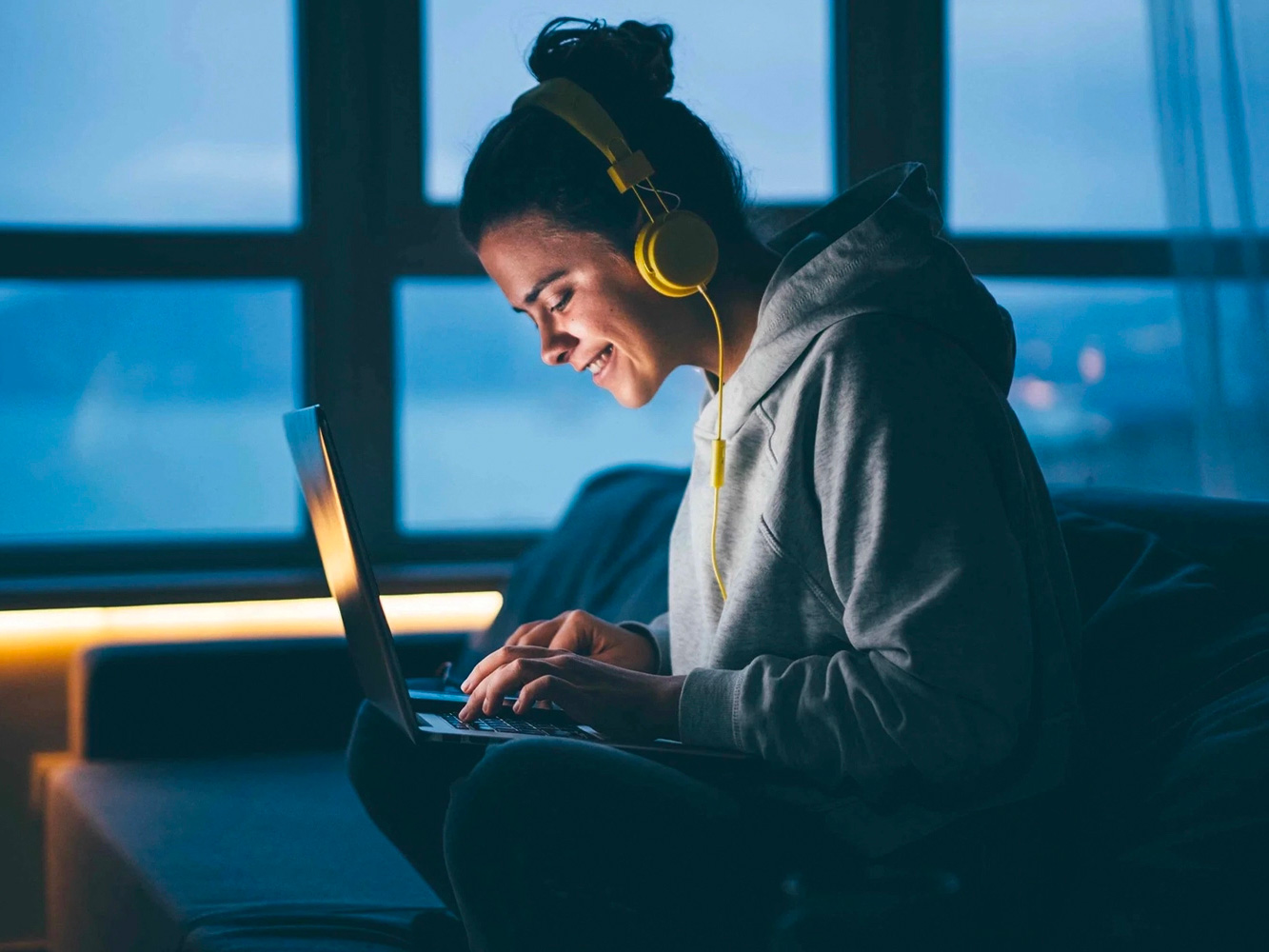 Una ragazza con le cuffie sul divano di casa, che utilizza felicemente il computer nella penombra.