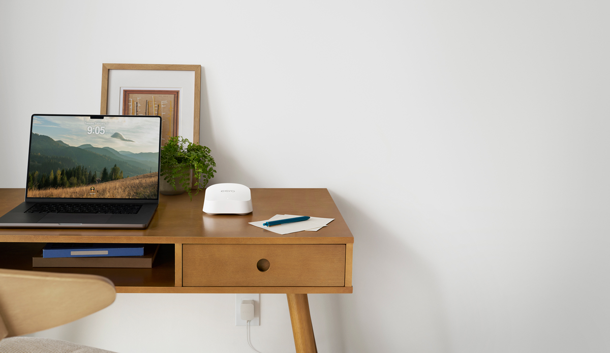 Una scrivania con sopra un computer, quadro, fogli e penna e l'Amazon eero 7, con la presa sotto alla scrivania che si intravede